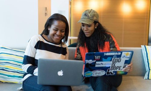 Two women working on their laptops while discussing their work